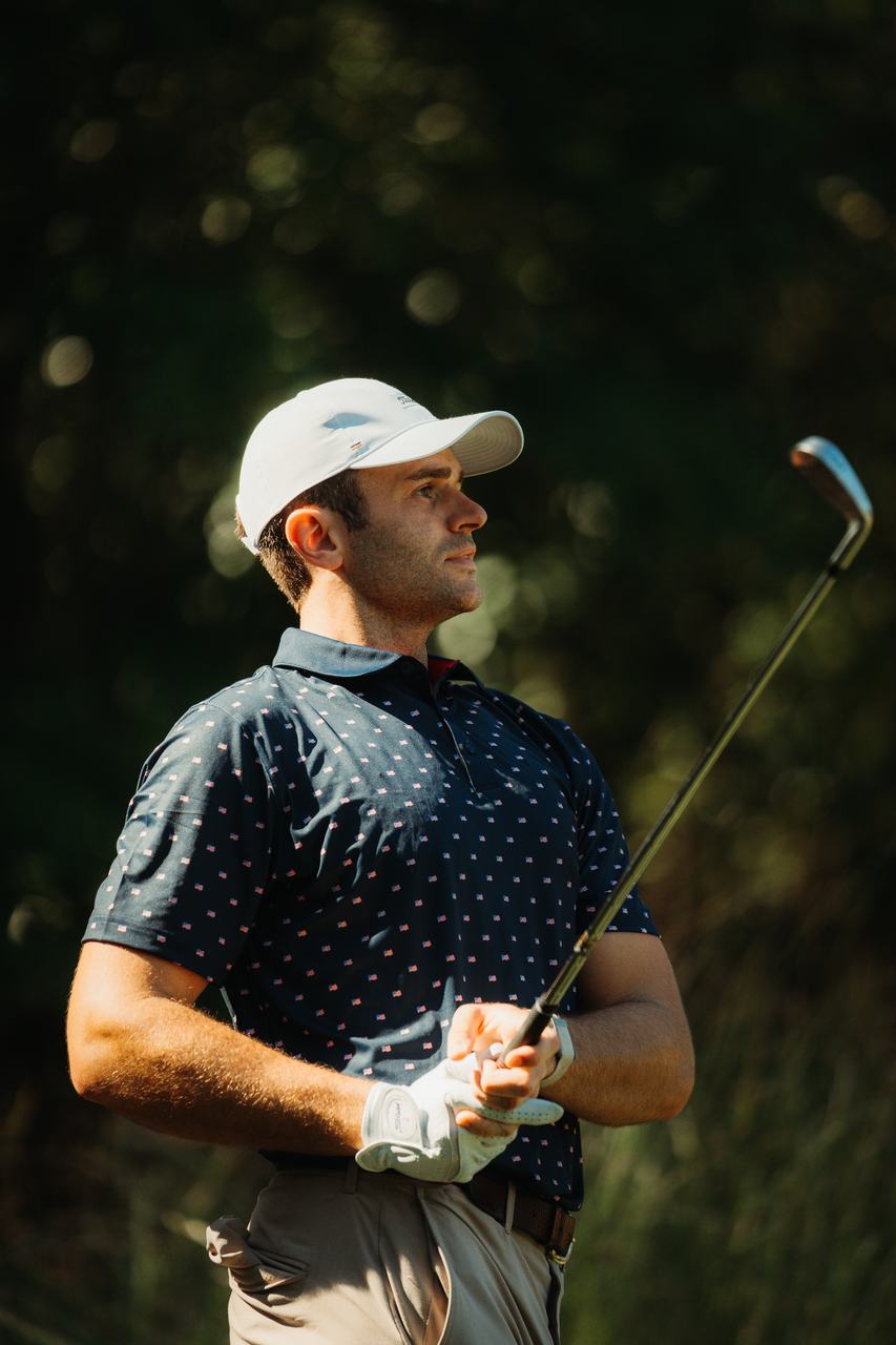 A golfer in a white cap and navy flag-print polo watches his shot, iron in hand, in warm sidelight