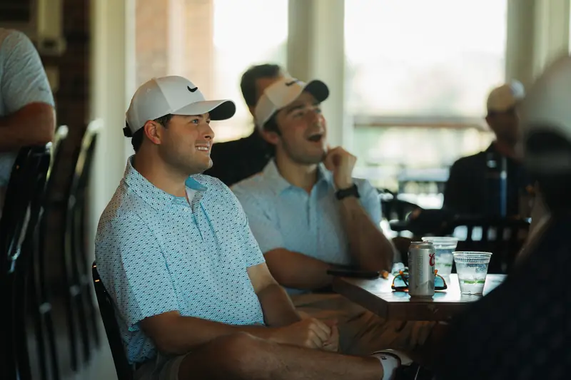 Two men in white caps and light blue polos laugh while watching something off-frame at a table with a can and cups, warm window light behind.