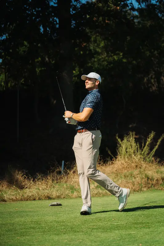 Golfer in navy dotted polo, khakis and grey cap watches his ball in a relaxed finish, warm sun on green fairway with dark trees behind.