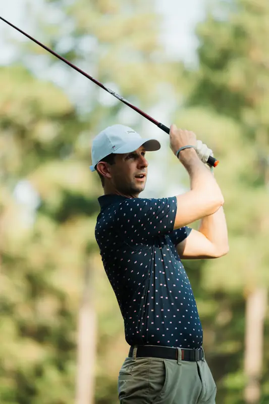 Close shot of a golfer in a light cap and navy polo following through, club streaking across the frame, creamy green background.