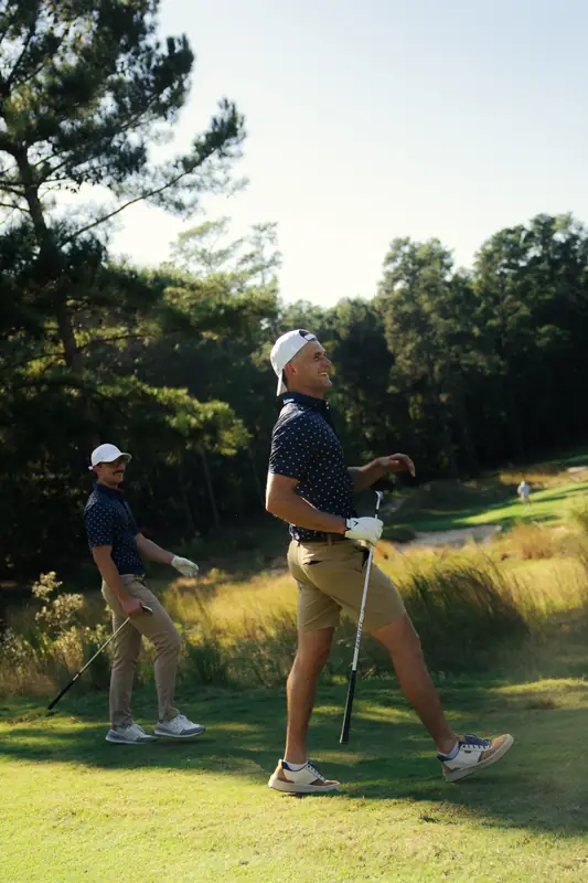 A tall golfer in navy polo, backwards cap and khakis laughs mid-stride on the fairway while a second golfer in a navy polo grins behind him, golden evening light and pines.