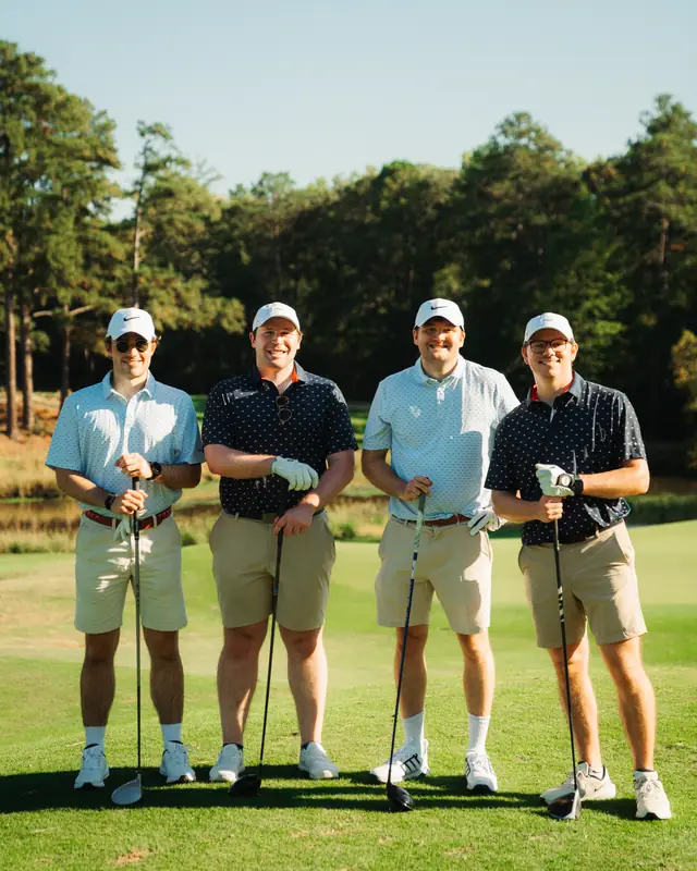 Four smiling golfers in white caps, alternating light blue and navy polos with khaki shorts, pose holding clubs on a sunlit fairway in front of pines.