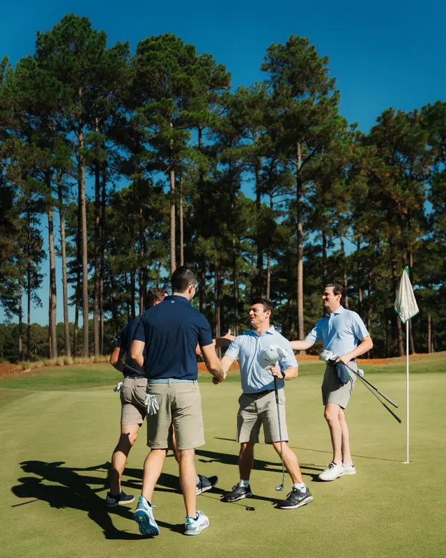 Four golfers in navy and light blue polos shake hands and smile on the green beside a flagstick under deep blue sky and pines.