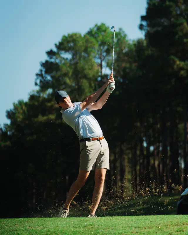 Golfer in light polo, khaki shorts, brown belt and dark cap in a full follow-through with club high, grass spraying, against blue sky and tall pines.
