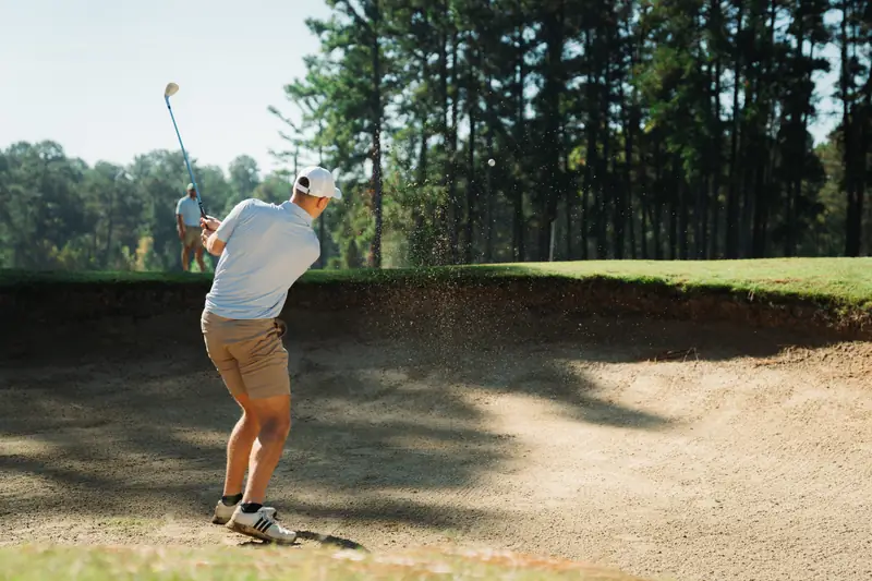 Golfer seen from behind in white cap and light polo blasts out of a sunny bunker, sand and ball in the air, a second golfer watching behind.