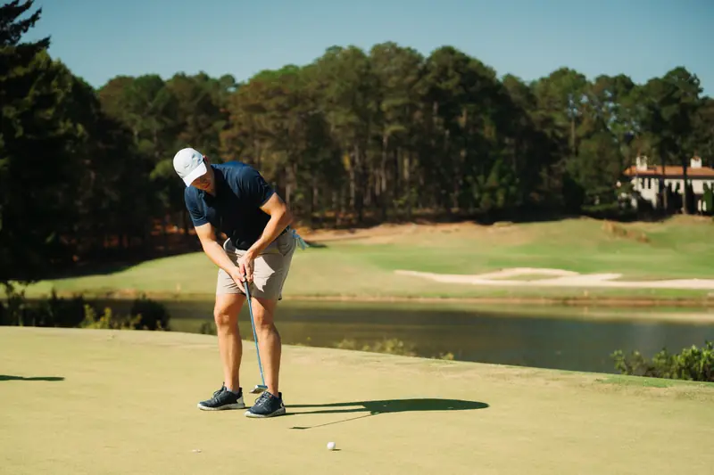 Wide shot of a golfer in white cap and navy polo putting on a sunlit green with a lake, bunkers and villa behind.