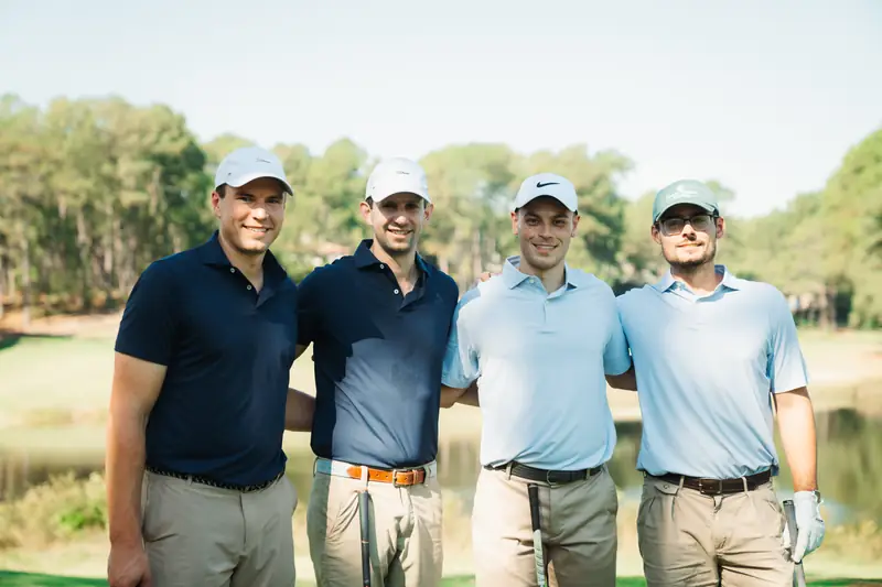 Landscape crop of four grinning golfers in caps and navy and light blue polos, arms linked, pond and pines behind.