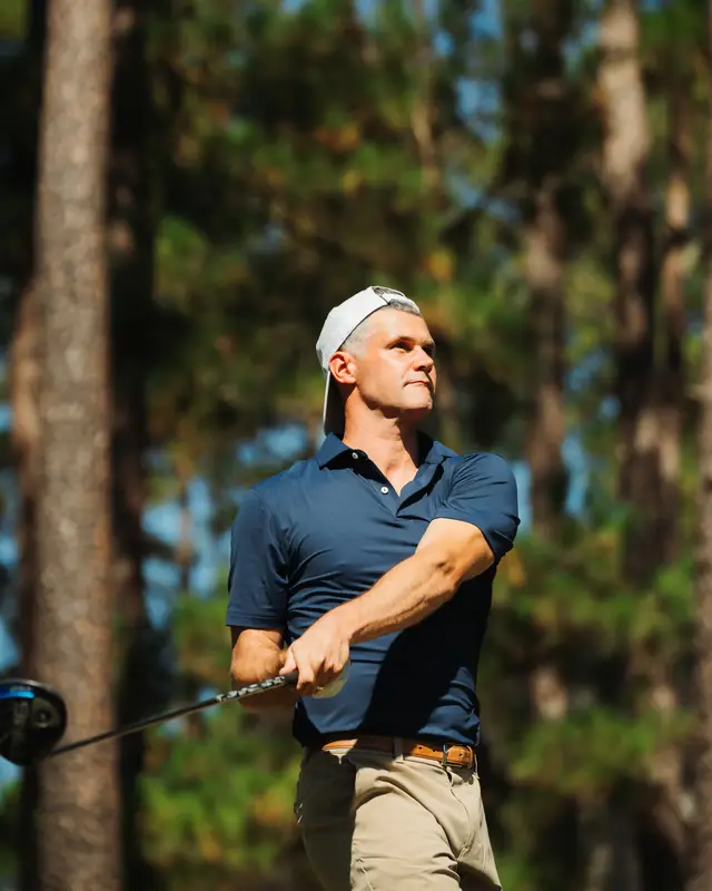 Same golfer in navy polo and backward cap in color, watching his shot with a strong follow-through among sunlit pines.