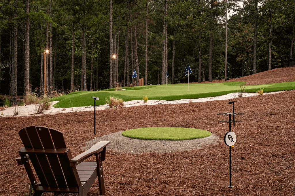 A wooden chair beside a tee box at The Aviary, looking up to a putting green with blue flags under tall pines